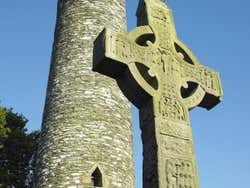 A stone statue of a cross in front of a stone tower under a blue sky